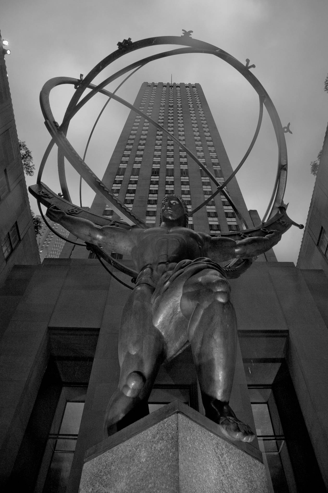 Statue of Atlas holding an armillary sphere in front of towering skyscraper, viewed from below in black and white.