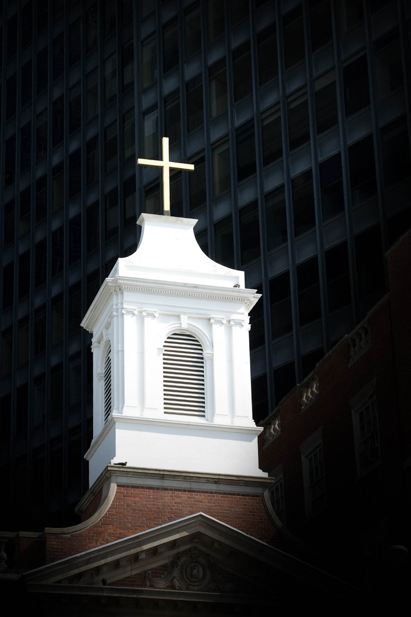 White church steeple with a gold cross against a dark, tall building backdrop.