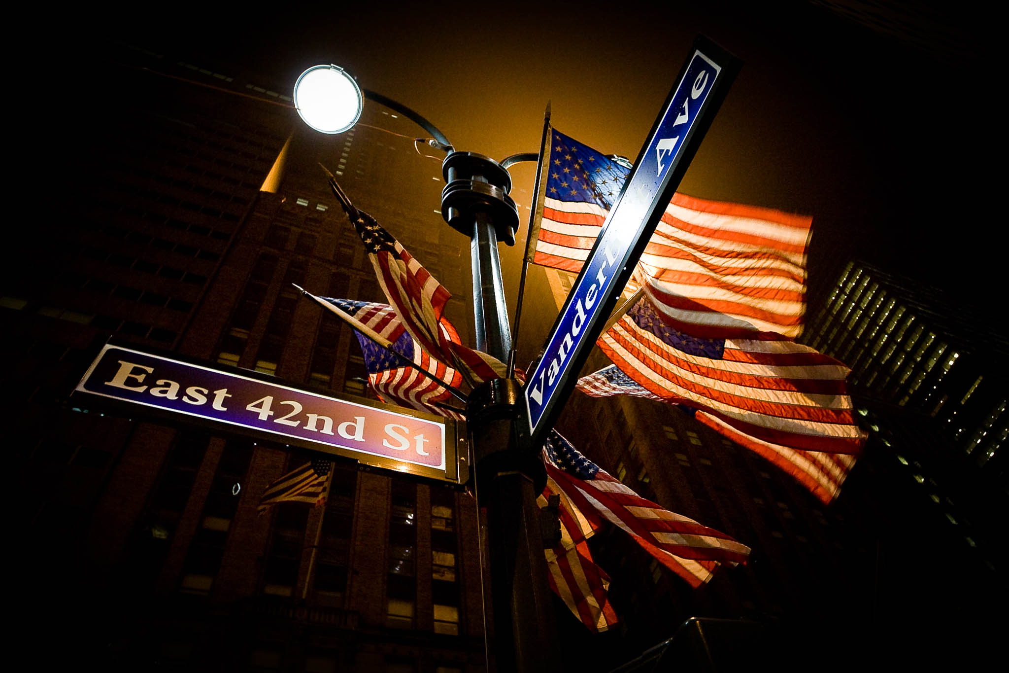Street signs at East 42nd St and Vanderbilt Ave with American flags illuminated at night.