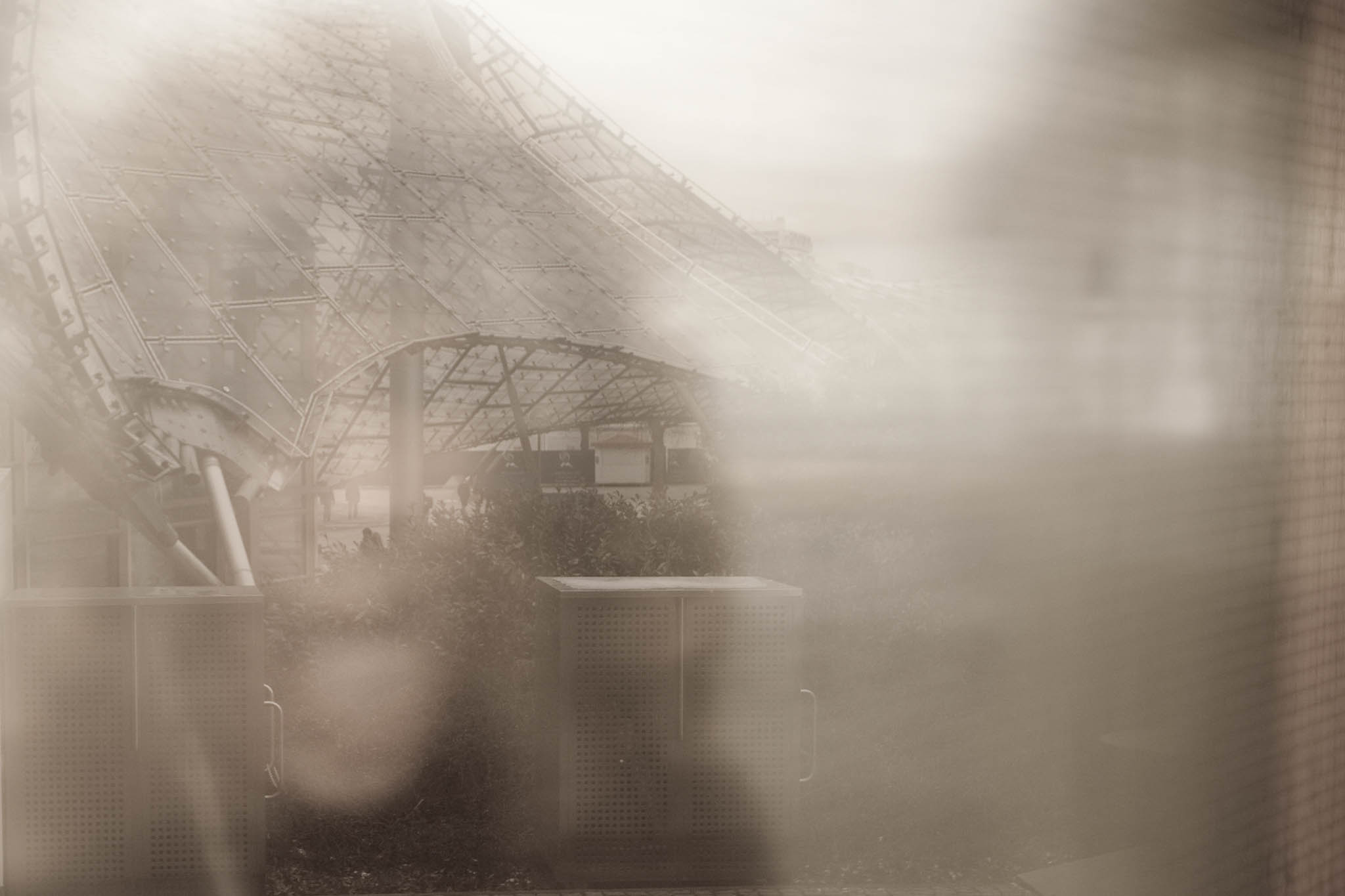 Blurred view of a geometric canopy structure with visible support beams and diffuse light, creating an abstract urban scene.