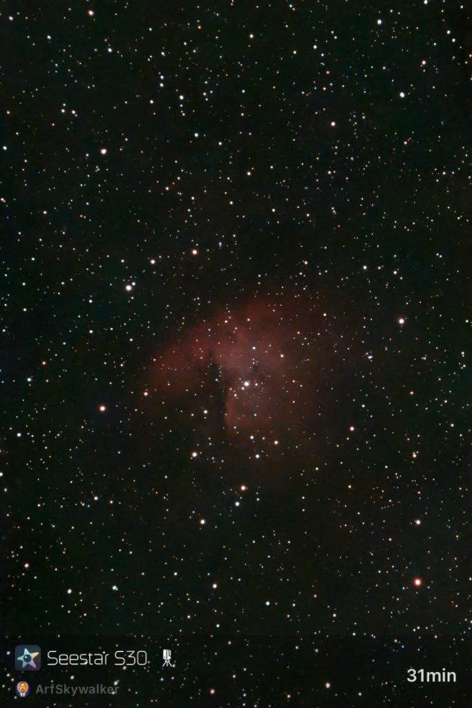 Starry sky with a dense field of stars, featuring a reddish nebula at the center amidst the dark space.