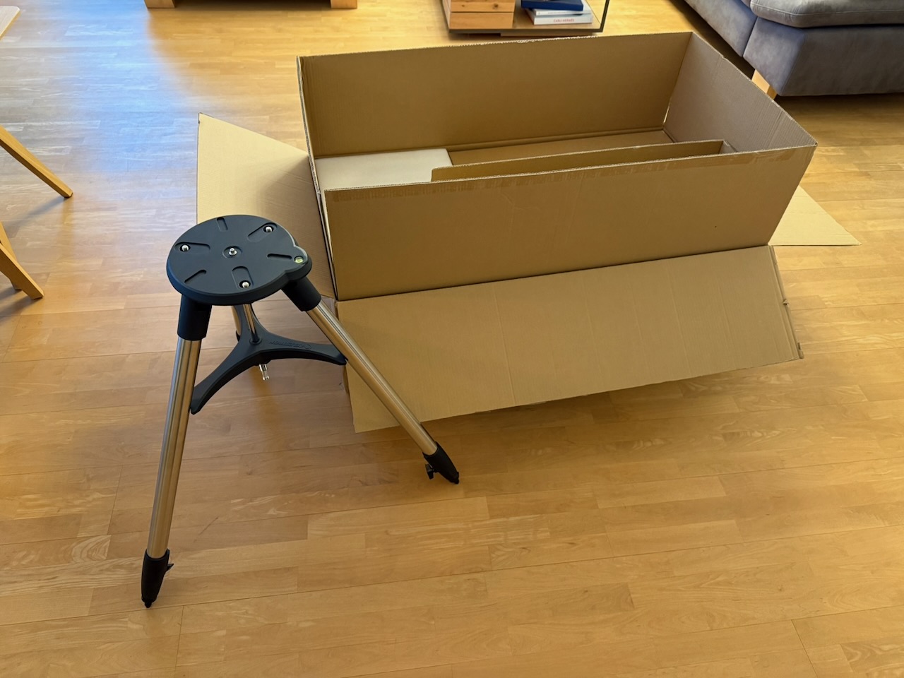 Large open cardboard box on a wooden floor beside a tripod stand in a living room setting.