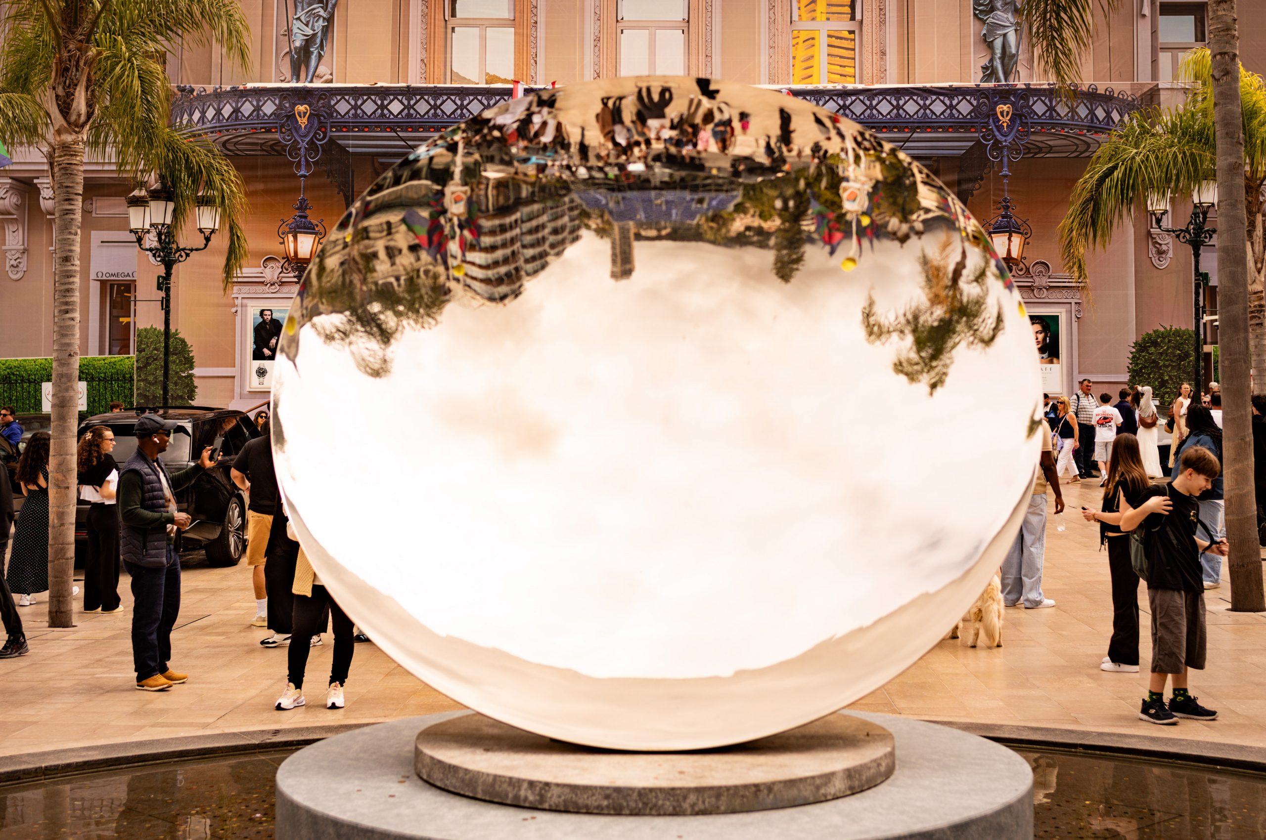 Large reflective sculpture in busy plaza with people and palm trees, building facade in the background.
