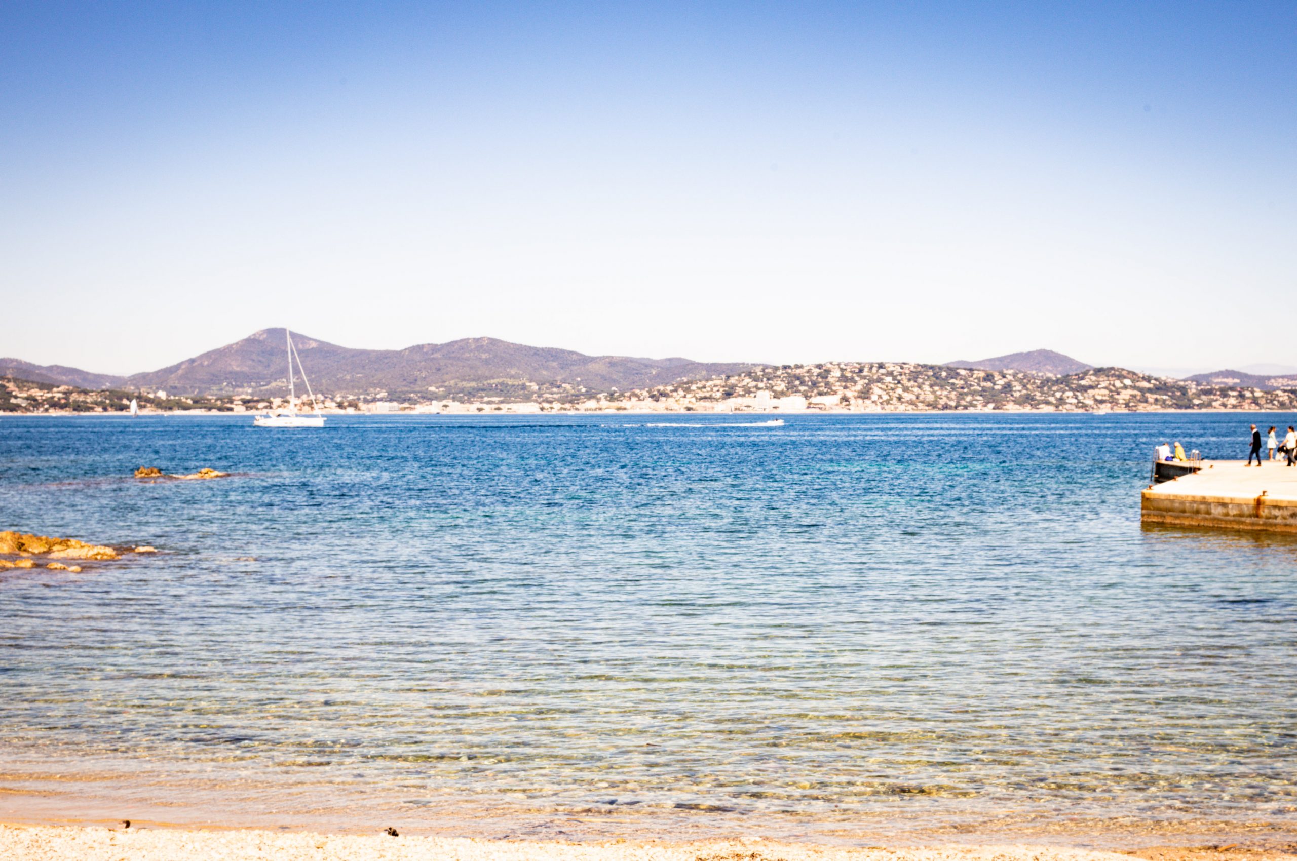 Clear blue water and distant mountains with a yacht on the sea; people standing on a pier under a bright sky.