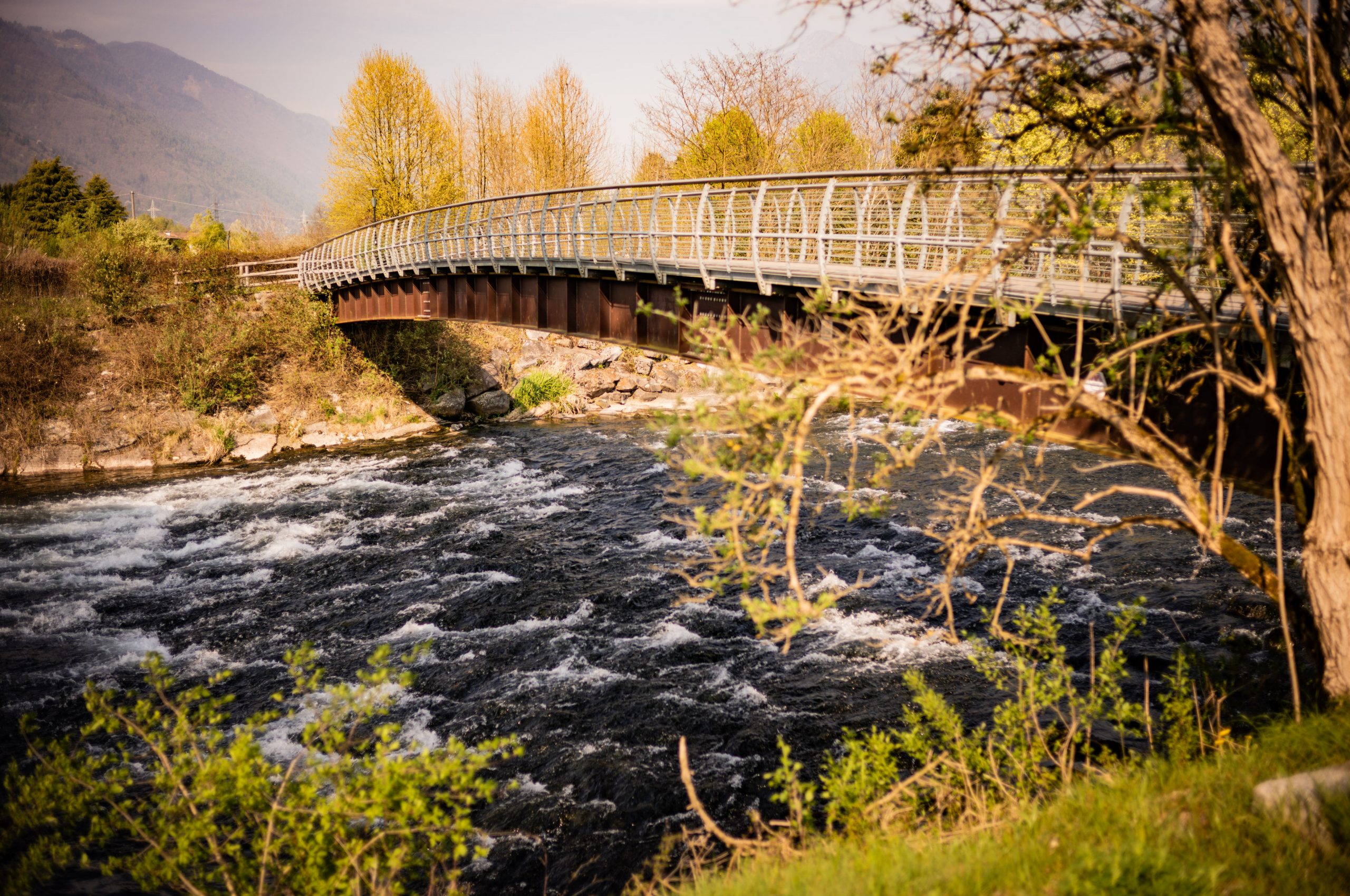 Bridge over a flowing river with lush greenery and mountains in the background, touched by warm sunlight.
