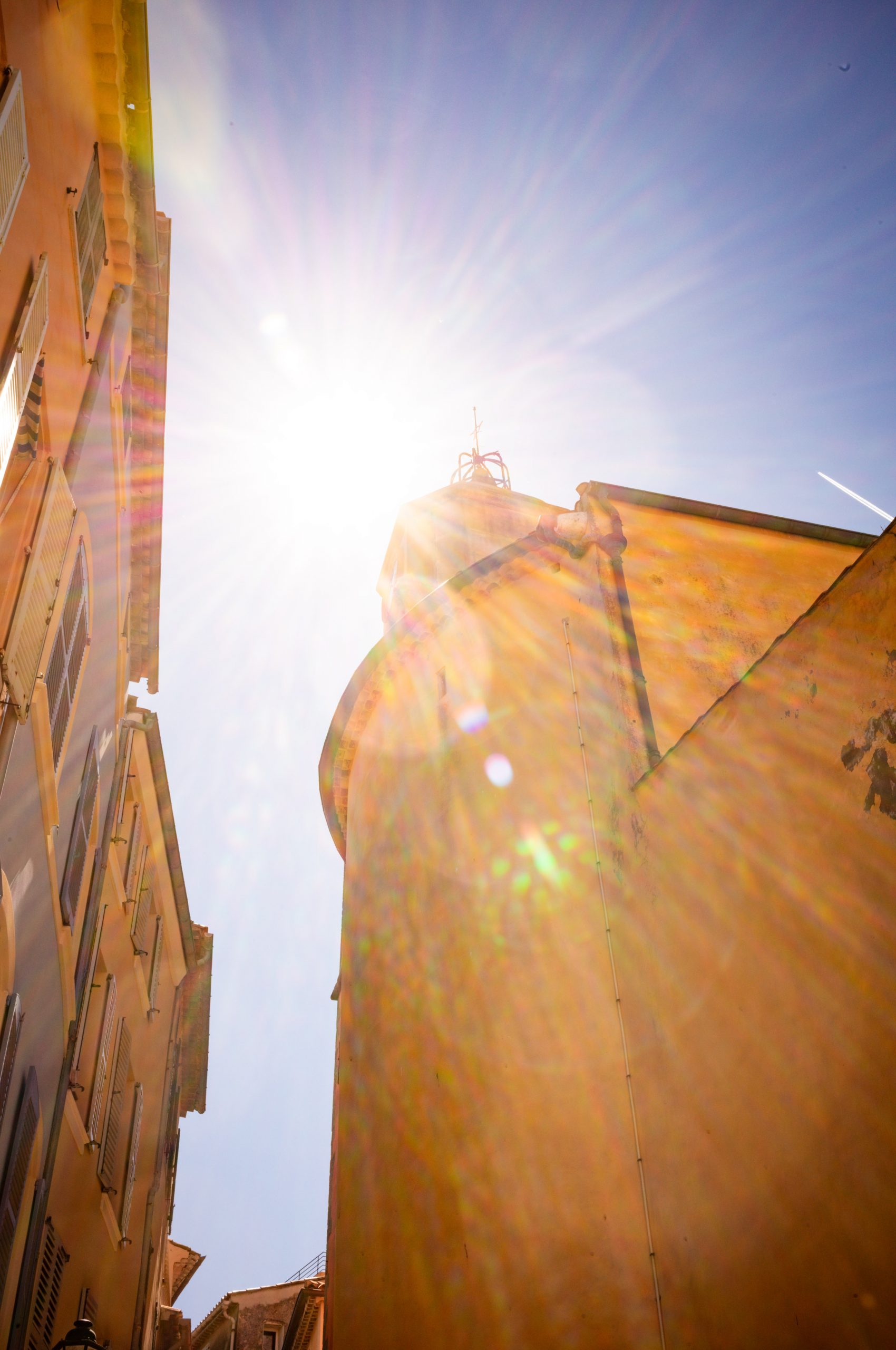 Sunlight bursts over old buildings in a narrow street, capturing vivid lens flares and a clear blue sky.