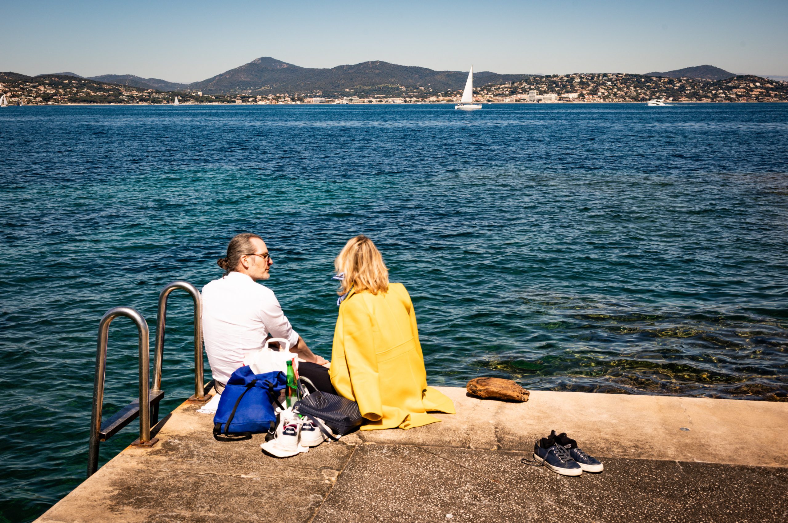 Couple sitting on a seaside pier by turquoise water, with a sailboat and distant mountains under a clear blue sky.