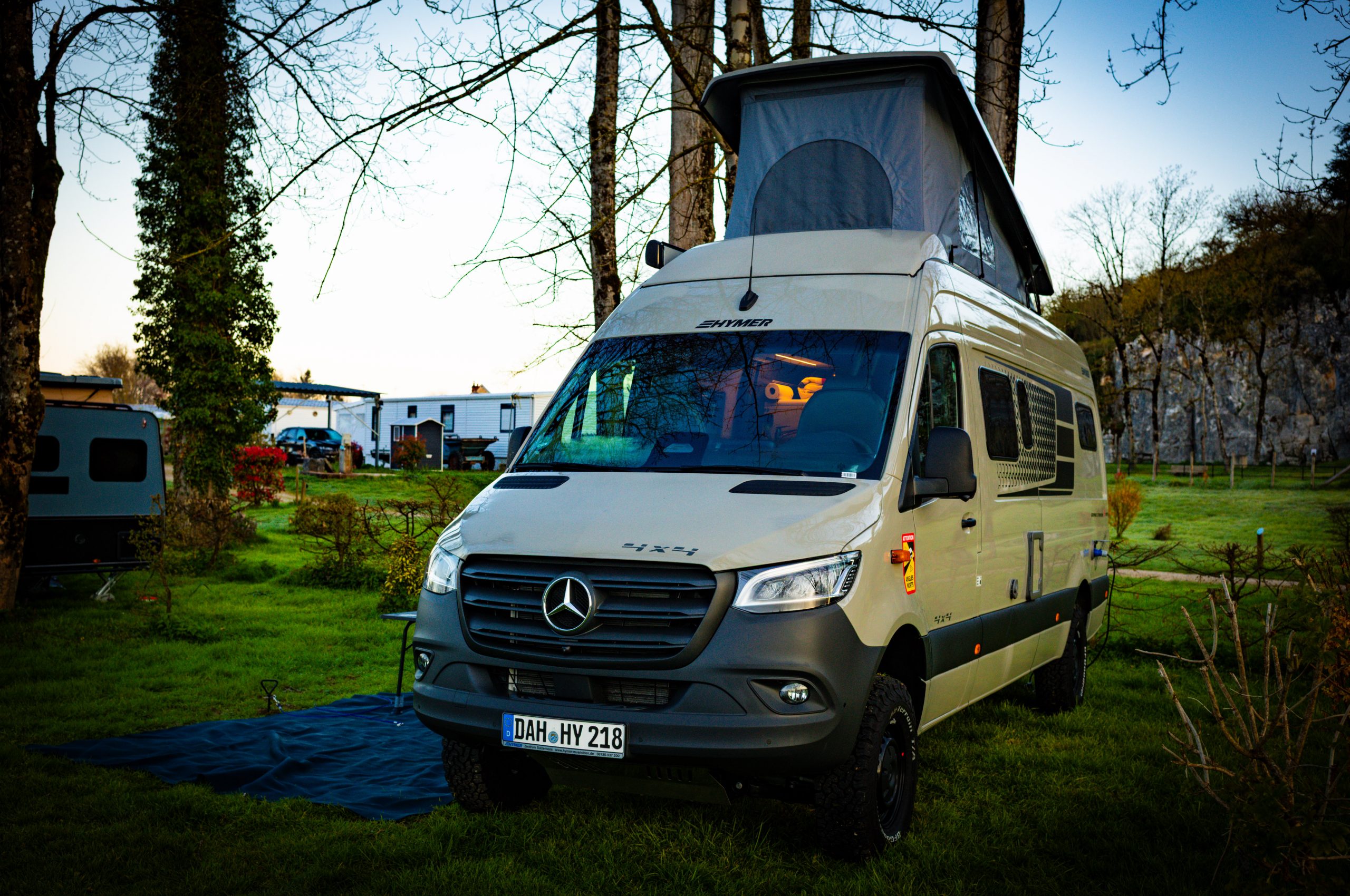 Hymer camper van parked in scenic campground with pop-top roof and trees, showcasing a serene outdoor travel setting.