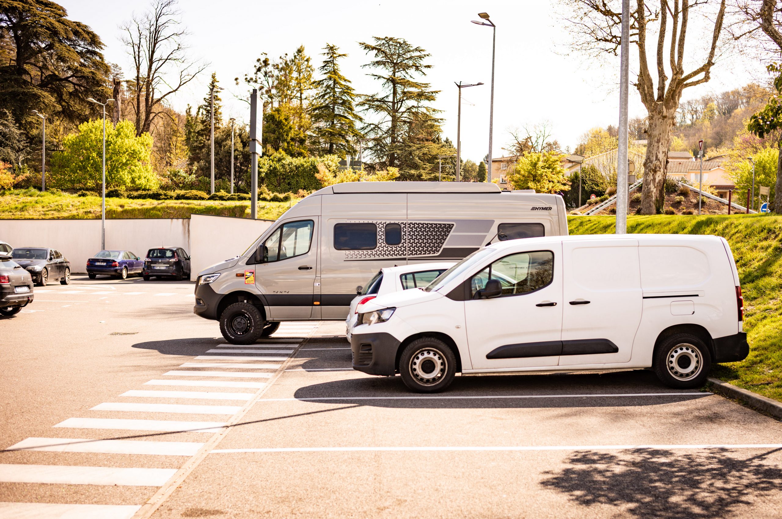 Camper van and white van parked in a sunny lot surrounded by greenery and trees.