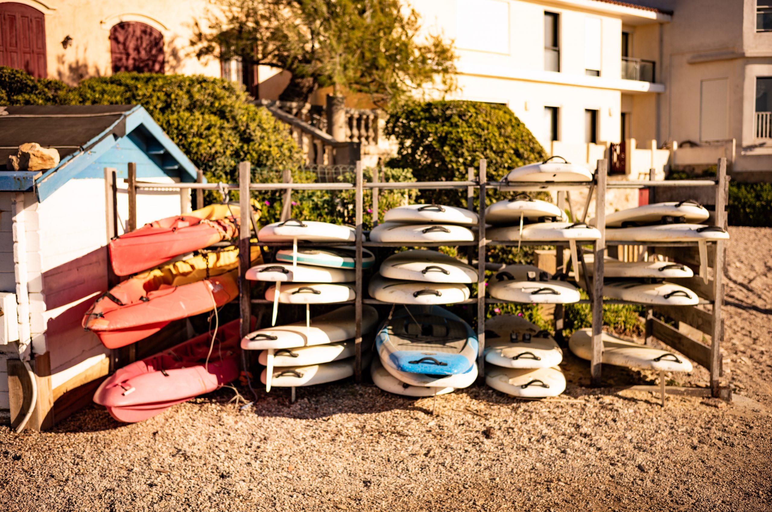 Stacked kayaks and paddleboards by a shed, set in a sunny, coastal storage area.