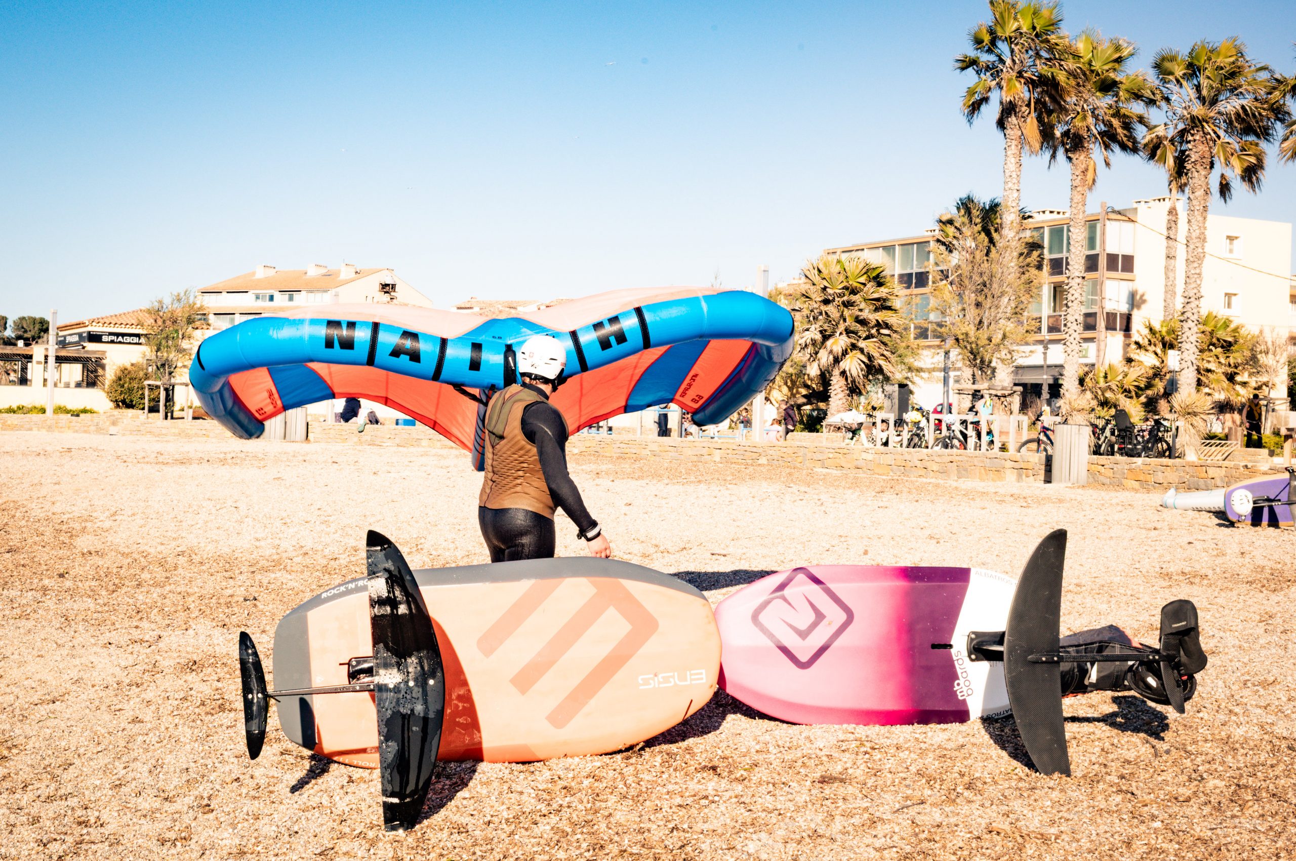 Person with a helmet holds a blue and orange kite next to hydrofoil boards on a sunny beach.