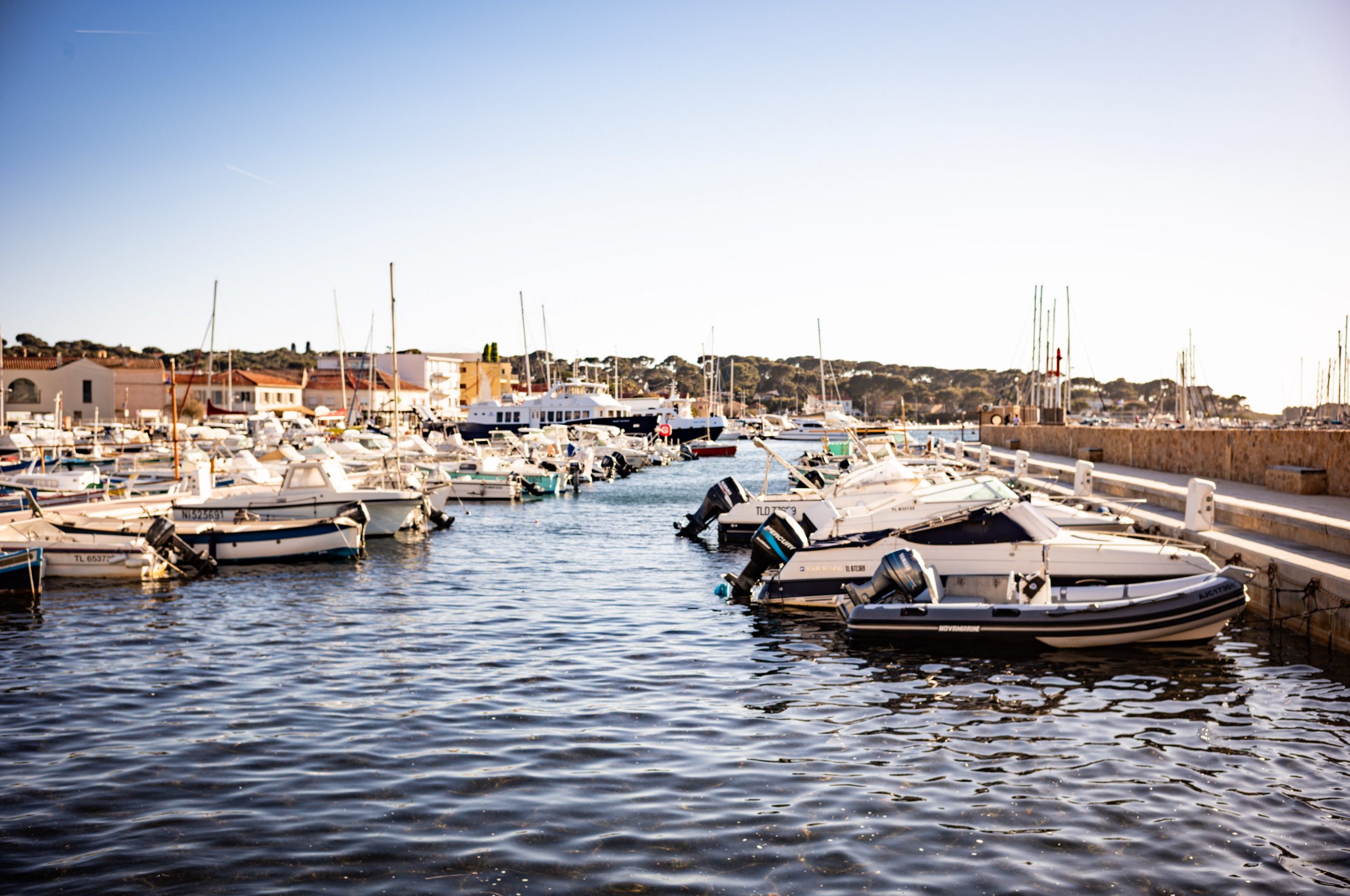 Sunny harbor with boats docked along a marina, clear blue sky, and distant hills in the background.