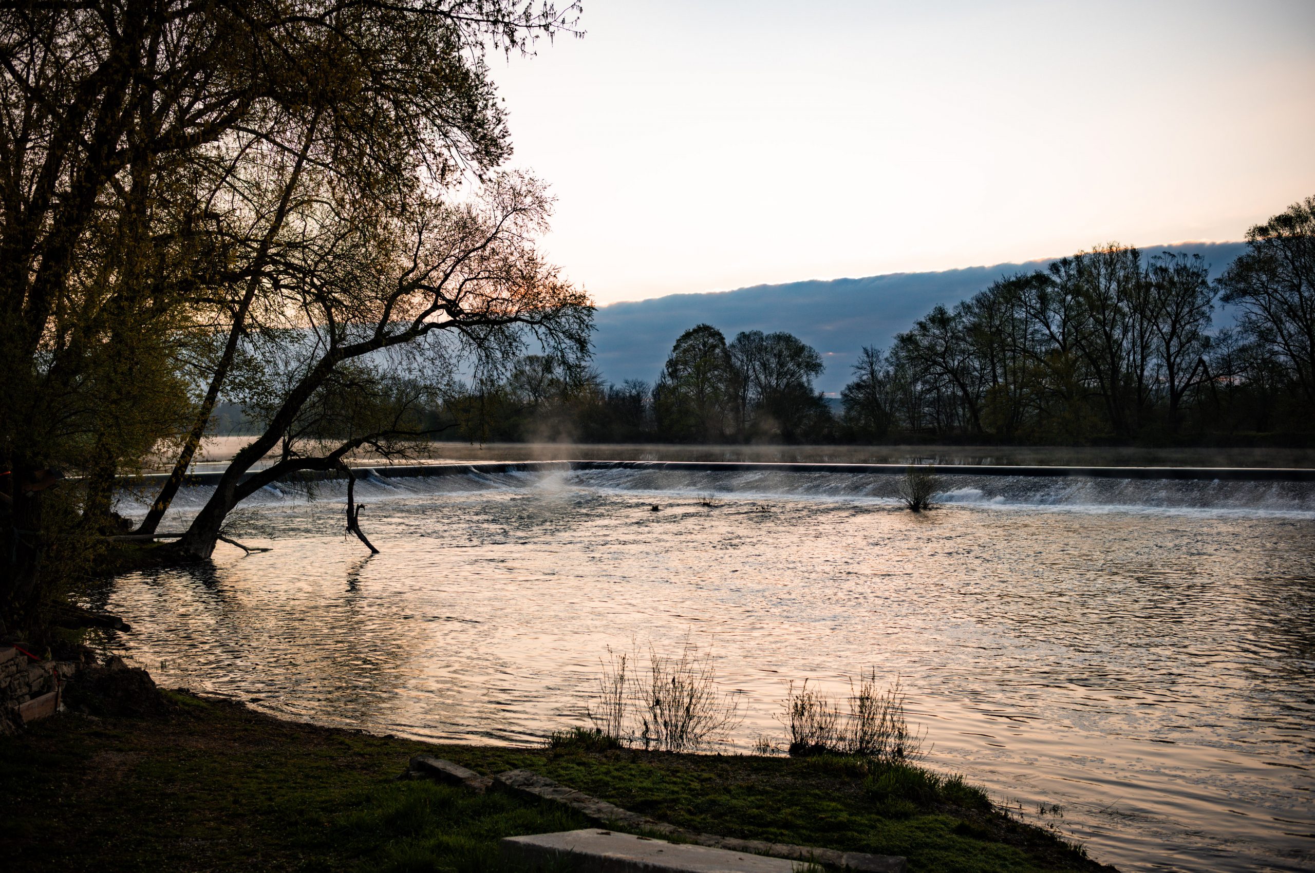 Tranquil river with trees at sunset, reflecting golden light and mist near a small waterfall.