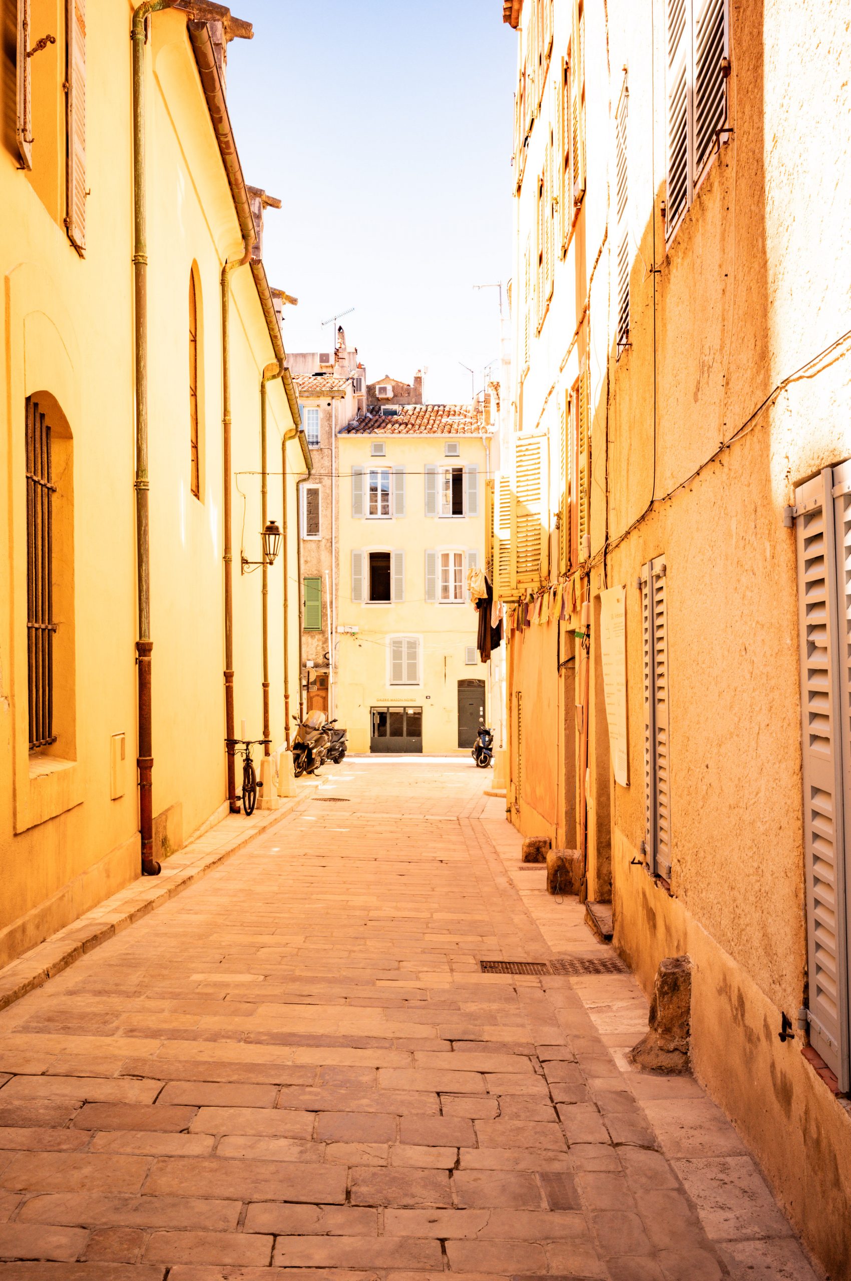 Sunlit narrow European alley with colorful facades, shutters, and scooters parked along cobblestone street.