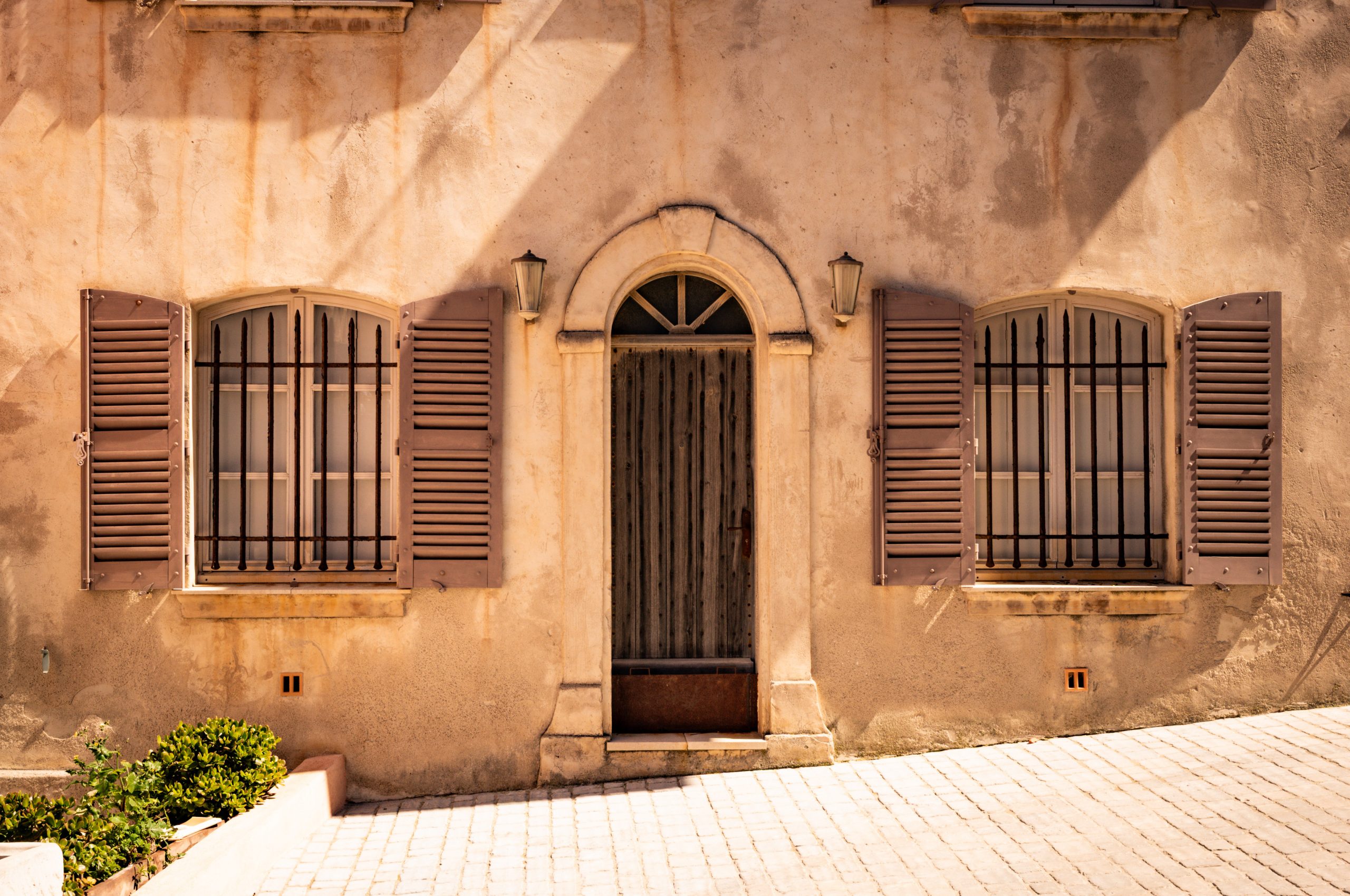 Vintage stucco facade with wooden shutters, arched door, and barred windows under warm sunlight.