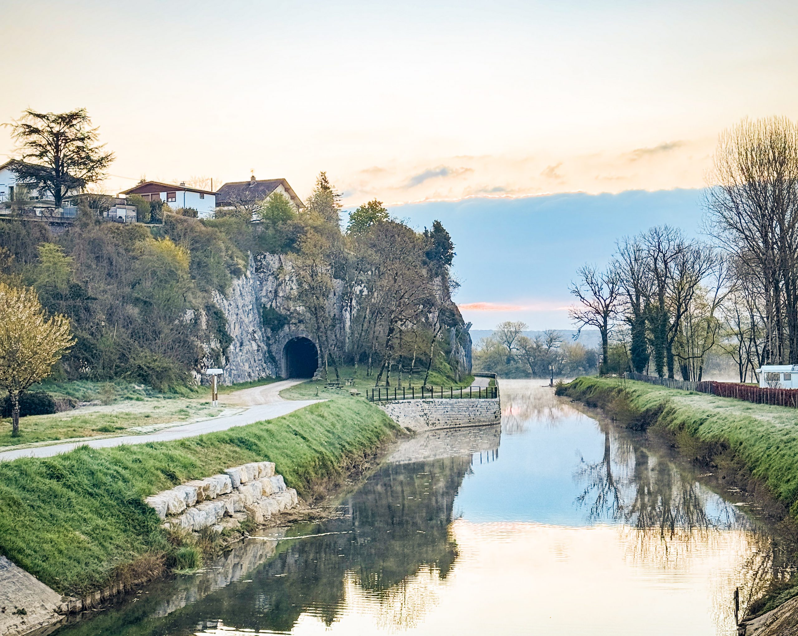 Picturesque canal at sunrise with a rocky hill, trees, and a path leading to a tunnel under a clear sky.