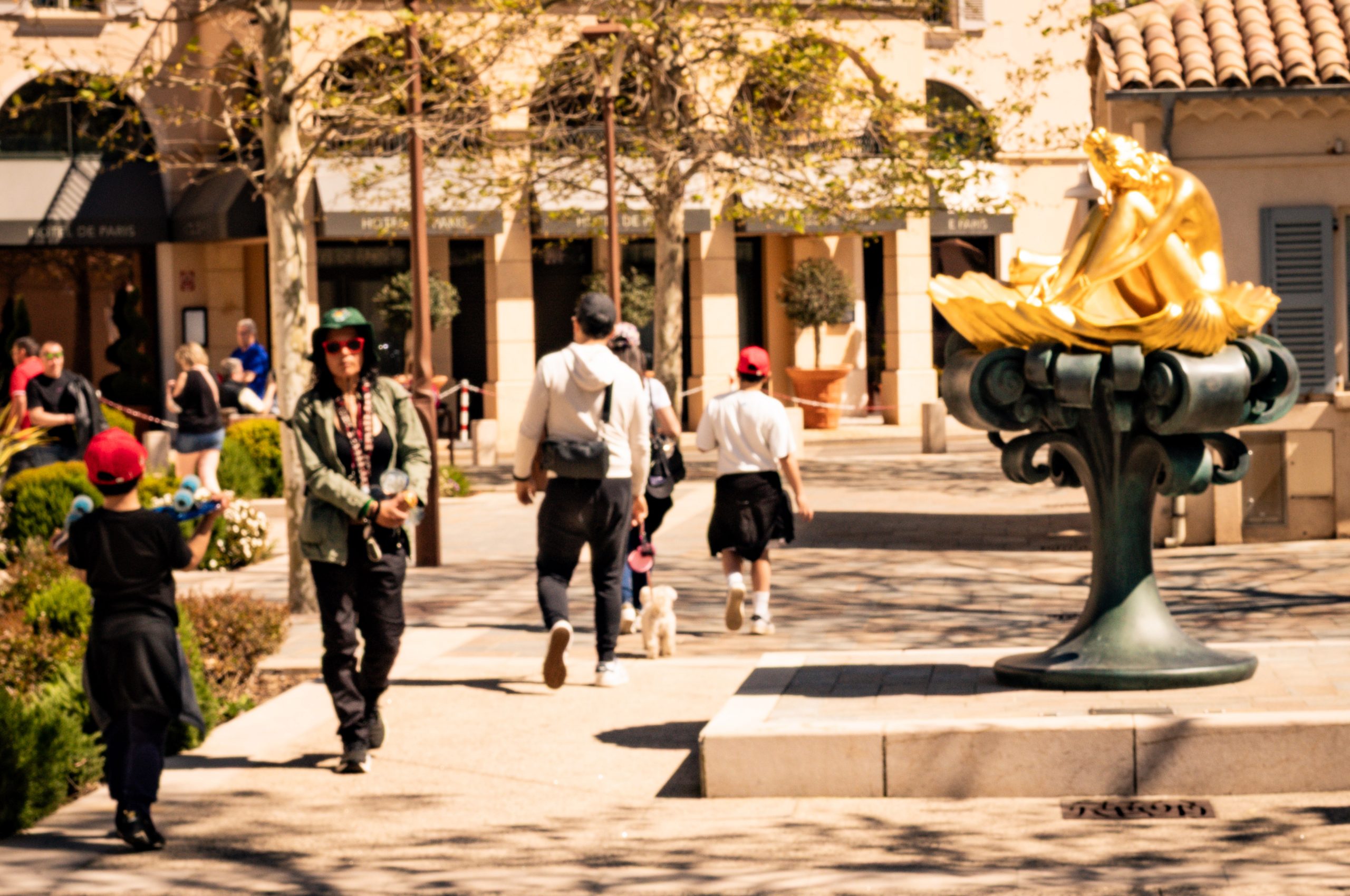 People walking in a sunny plaza with a golden statue, trees, and a Mediterranean-style building in the background.