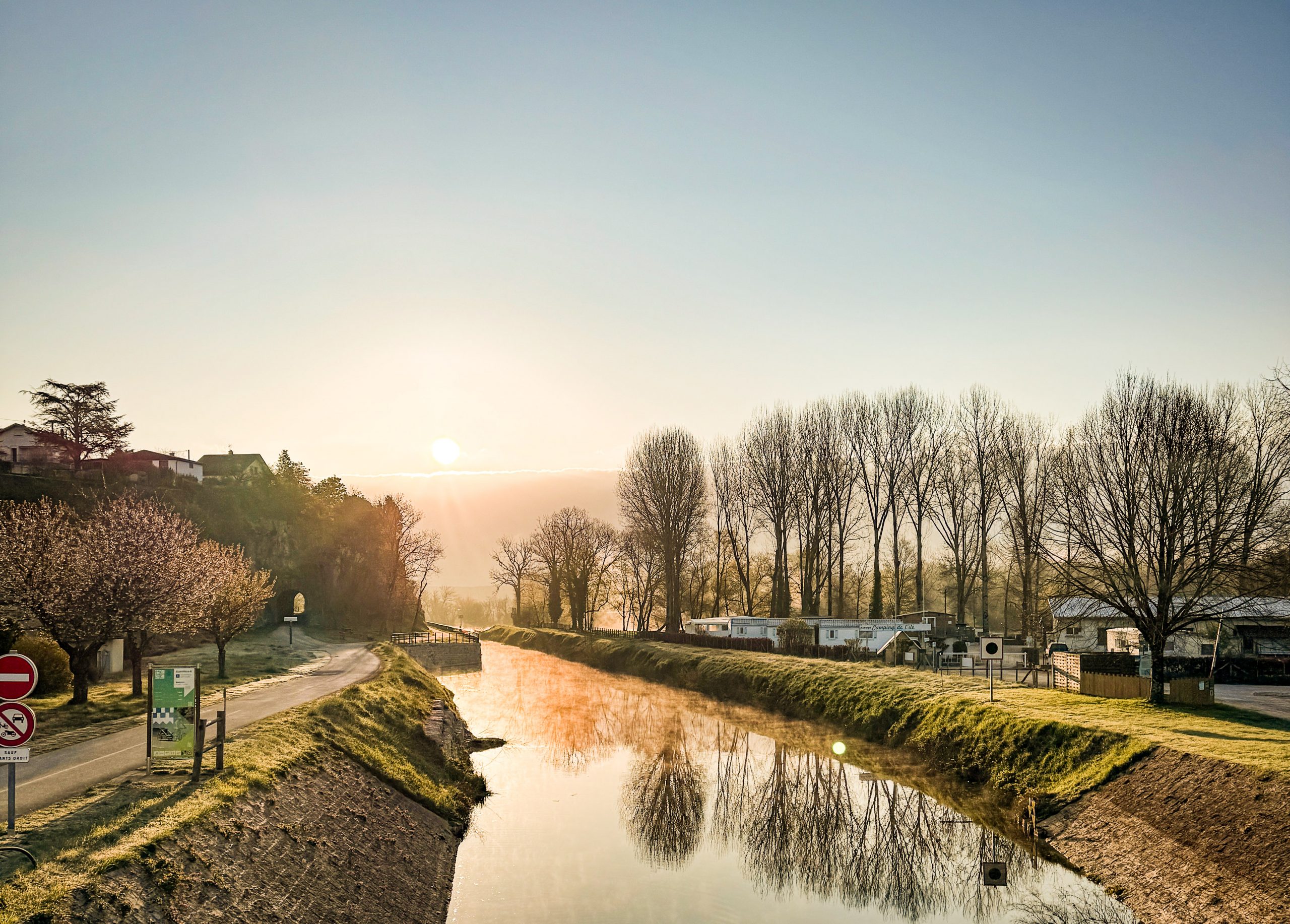 Sunrise over tranquil canal with reflections, tree-lined banks, and distant houses create a serene landscape.