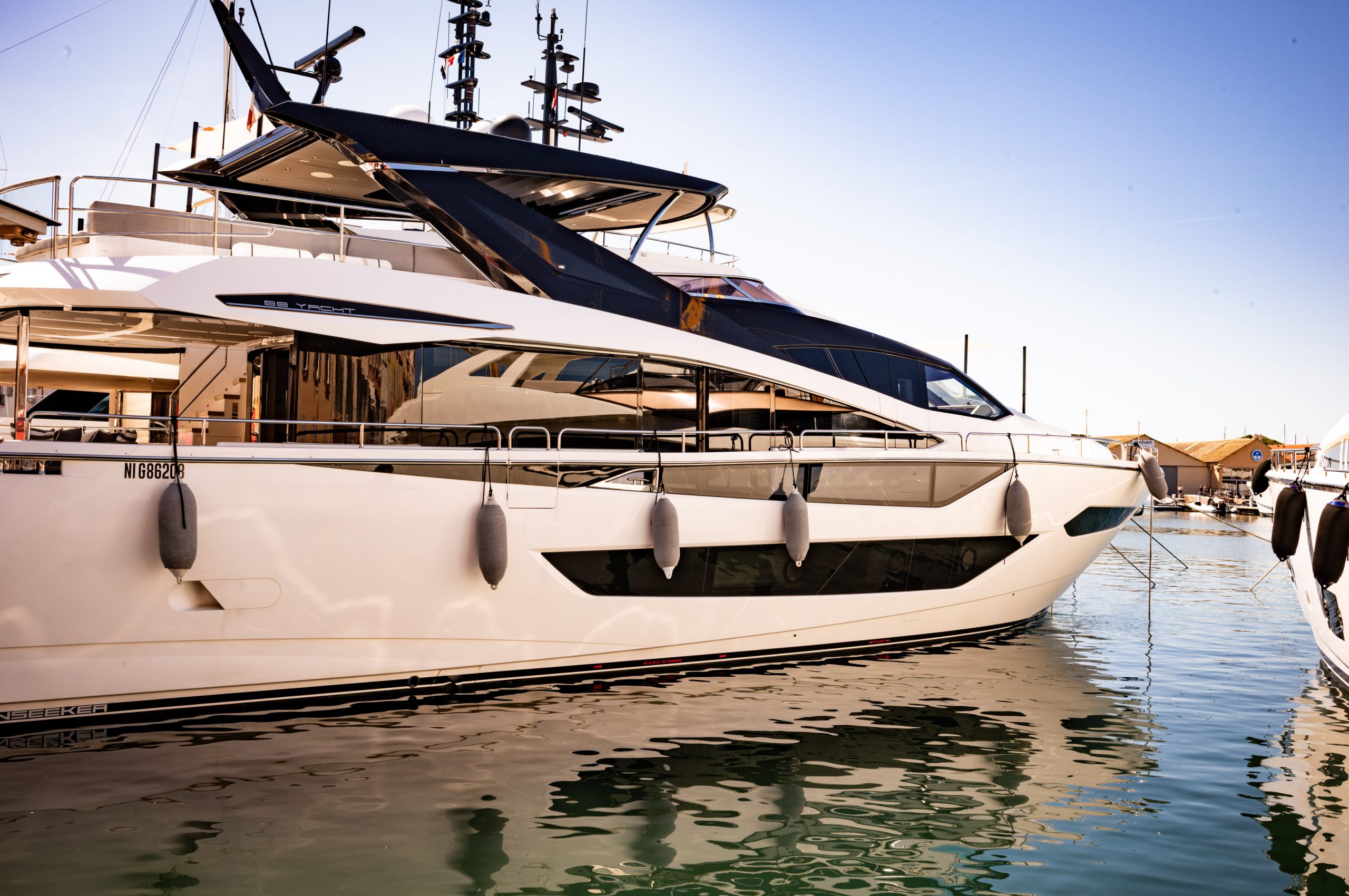 Luxury yacht docked in marina with sleek design and modern features, reflecting on calm water under clear blue sky.