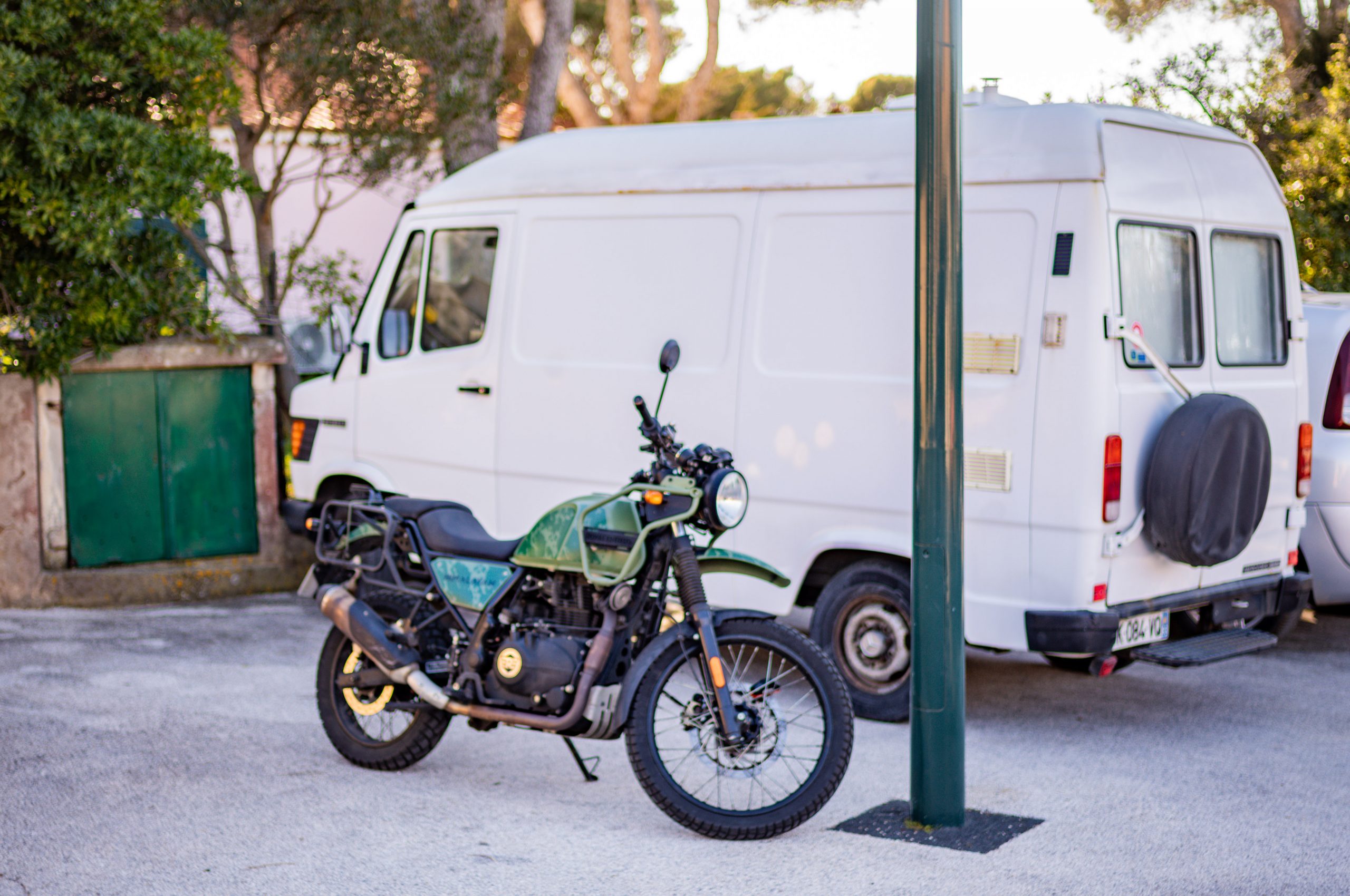 Green motorcycle parked in front of a white van on a sunny street, surrounded by trees.